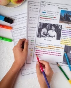 Hands holding a pencil writing on a passage about helen keller with common core questions on papers behind the passage