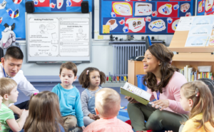 A teachers reads to her class while sitting on the floor in a circle. An open book is in her hand, and she is looking directly at a student while discussing predicting strategies with the group.